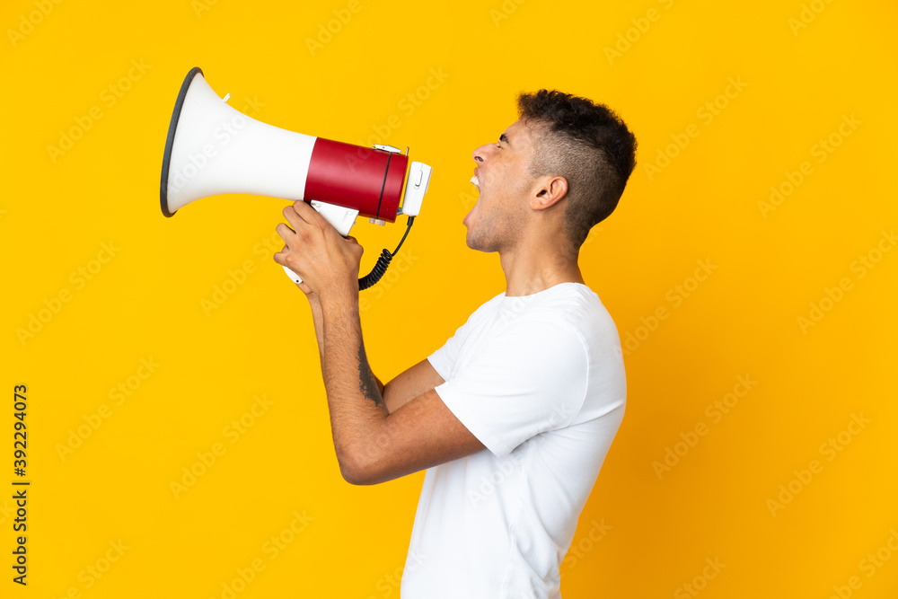 Young brazilian man isolated on yellow background shouting through a megaphone to announce something in lateral position Young brazilian man isolated on yellow background shouting through a megaphone to announce something in lateral position