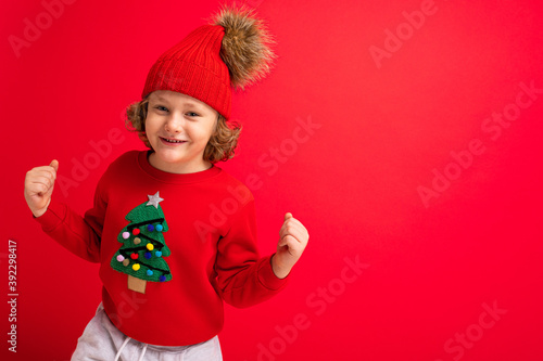 cool blond kid in warm hat and sweater with christmas tree on red background fooling around, christmas concept