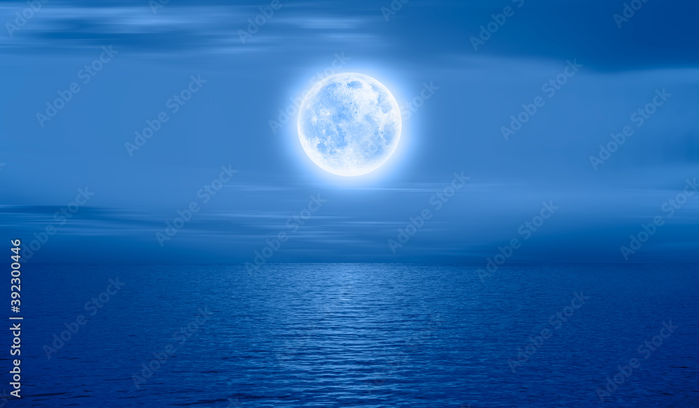 Night sky with full bright moon in the clouds, blue sea in the foreground 