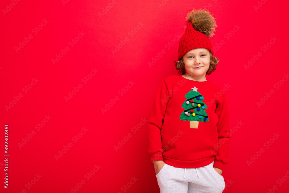 cheerful funny boy on a red background in a warm hat and sweater with a Christmas tree
