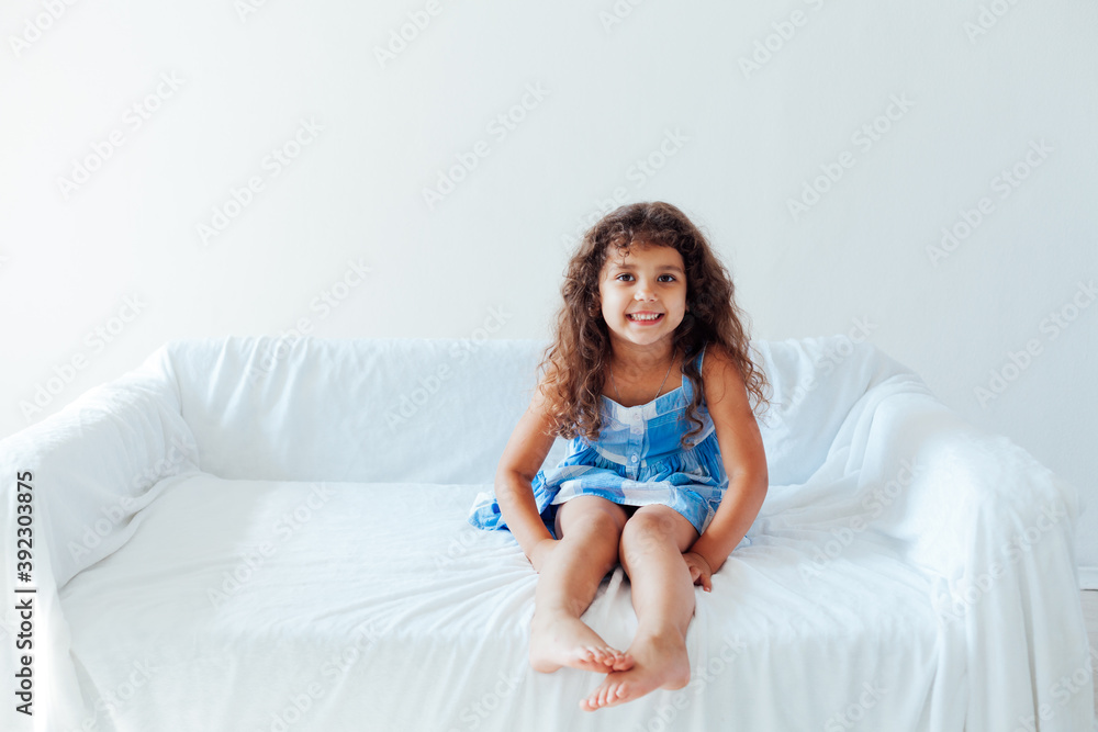 Beautiful little curly girl in a blue dress on the white couch in the room Beautiful little curly girl in a blue dress on the white couch in the room