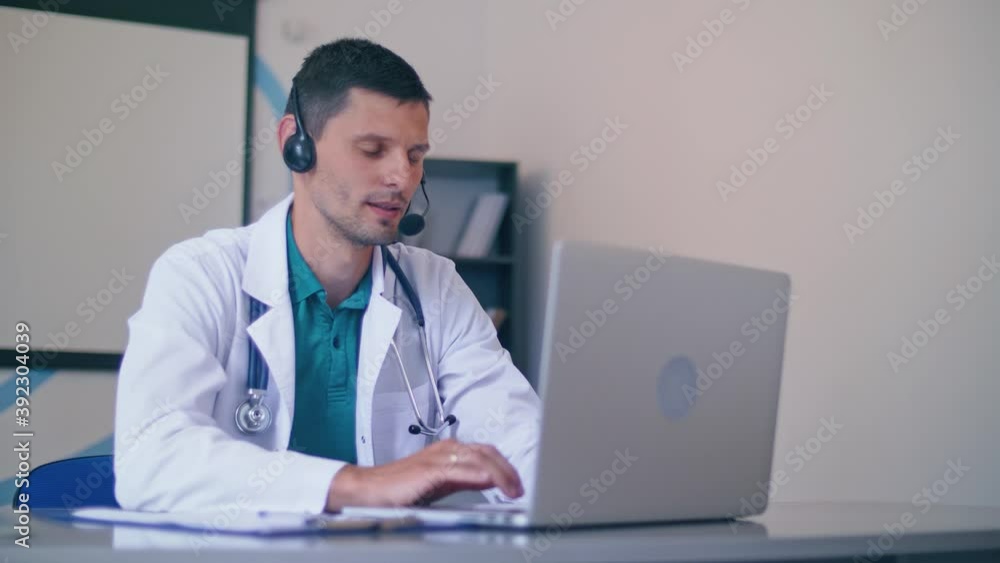 Friendly Male Doctor in White Medical Coat With Headphones Making Conference Call on Laptop.Remote Consulting Patient Online From Healthcare Hospital. Telemedicine. Friendly Male Doctor in White Medical Coat With Headphones Making Conference Call on Laptop.Remote Consulting Patient Online From Healthcare Hospital. Telemedicine.