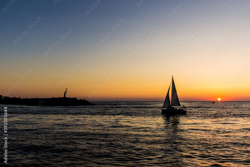 Silhouette of a sailboat on the sea next to a dock with a sunset on the horizon.