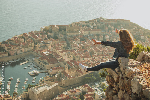 Happy excited girl sitting on the edge of a rock with the old city of Dubrovnik behind her. Beautiful ancient city glowing during an afternoon sunset, visiting during the covid season