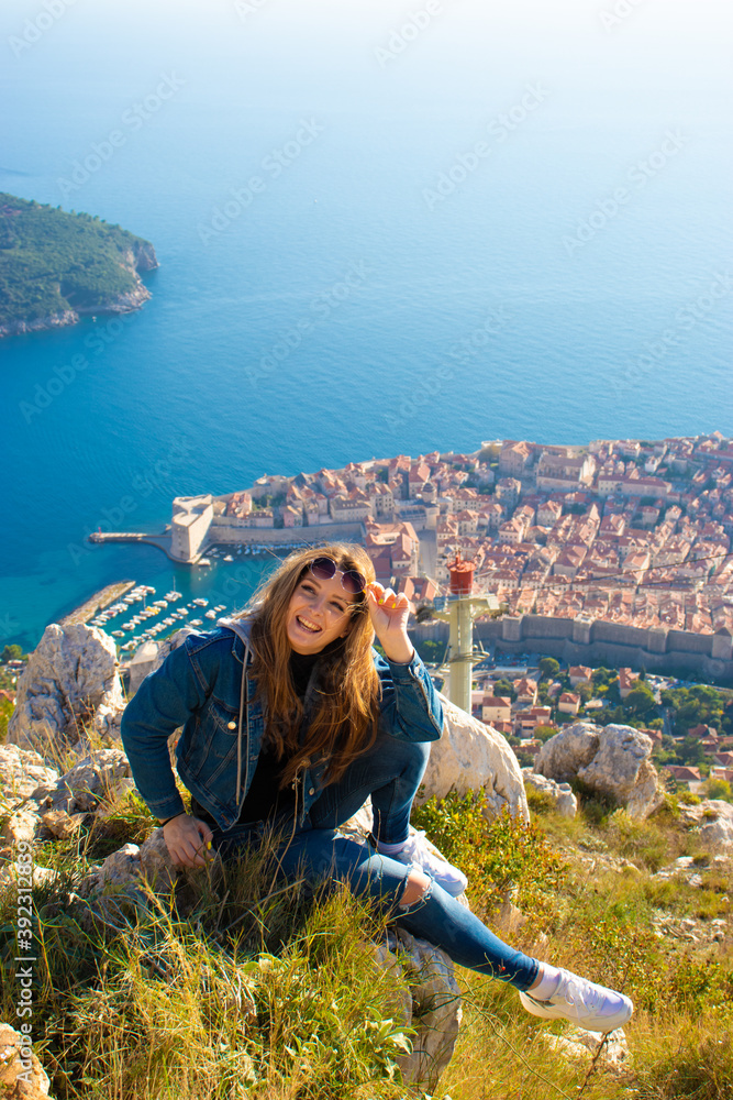Naklejka premium Vertical shot of a brunette sitting on the stones on the Srd mountain above the city of Dubrovnik, posing for a photo and laughing as she holds her sunglasses
