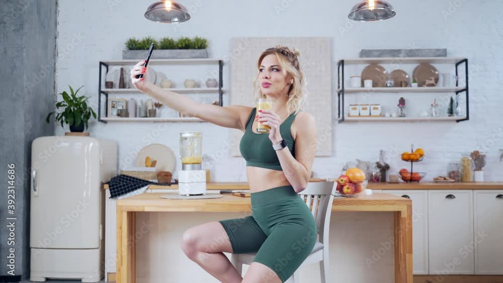 fitness young woman posing for photo with glass of fresh vegetable fruit juice in kitchen interior. Happy woman drinking healthy drink in front of camera phone. Pretty girl making video on smartphone fitness young woman posing for photo with glass of fresh vegetable fruit juice in kitchen interior. Happy woman drinking healthy drink in front of camera phone. Pretty girl making video on smartphone