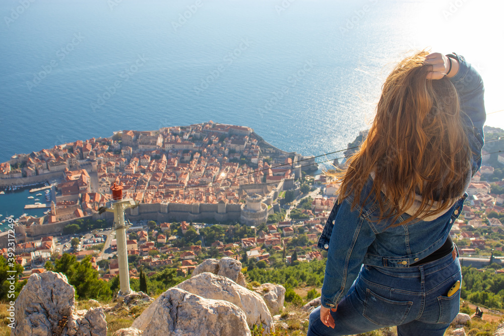 Over the shoulder view of a girl standing on top of the Srd mountain, looking down on Dubrovnik city in the distance. Ancient famous city surrounded by the bright adriatic sea