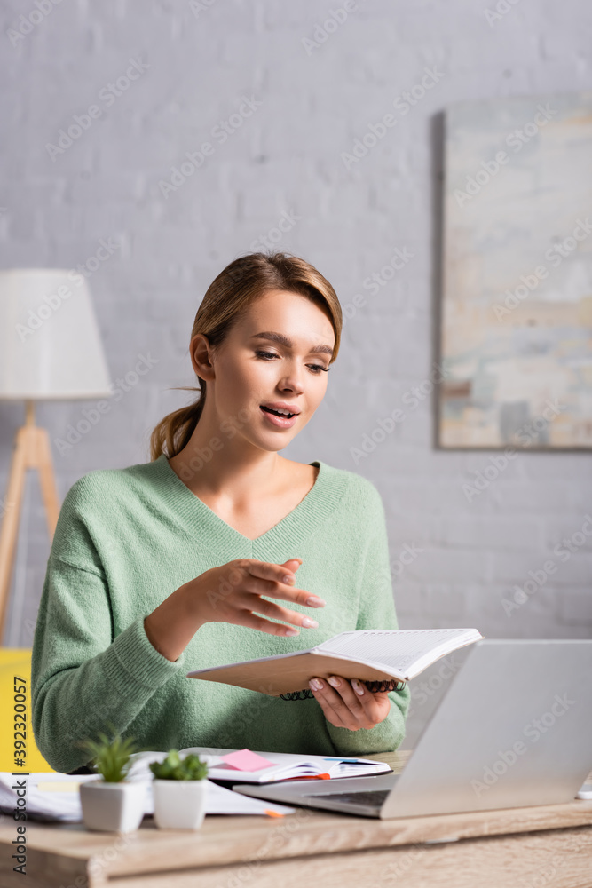 Smiling freelancer holding notebook during video call on laptop on blurred foreground