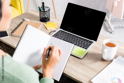 Laptop with blank screen near cup of tea and papers on table near freelancer writing on notebook on blurred foreground