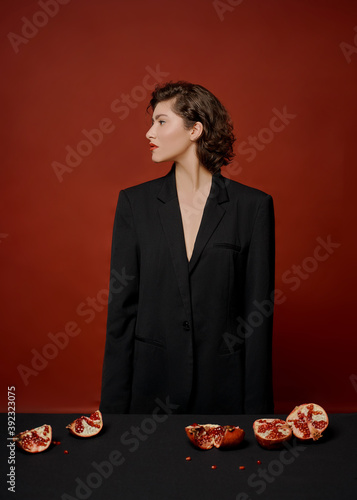 a beautiful young woman in a black jacket is sitting at a table on a red background. holds halves and pieces of pomegranate fruit
