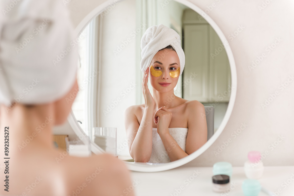 A young woman in white clothes and with a towel on her head looks in the mirror and applies gold plasters under her eyes, taking care of her skin in the morning. Woman applying gold spots on her face. A young woman in white clothes and with a towel on her head looks in the mirror and applies gold plasters under her eyes, taking care of her skin in the morning. Woman applying gold spots on her face.