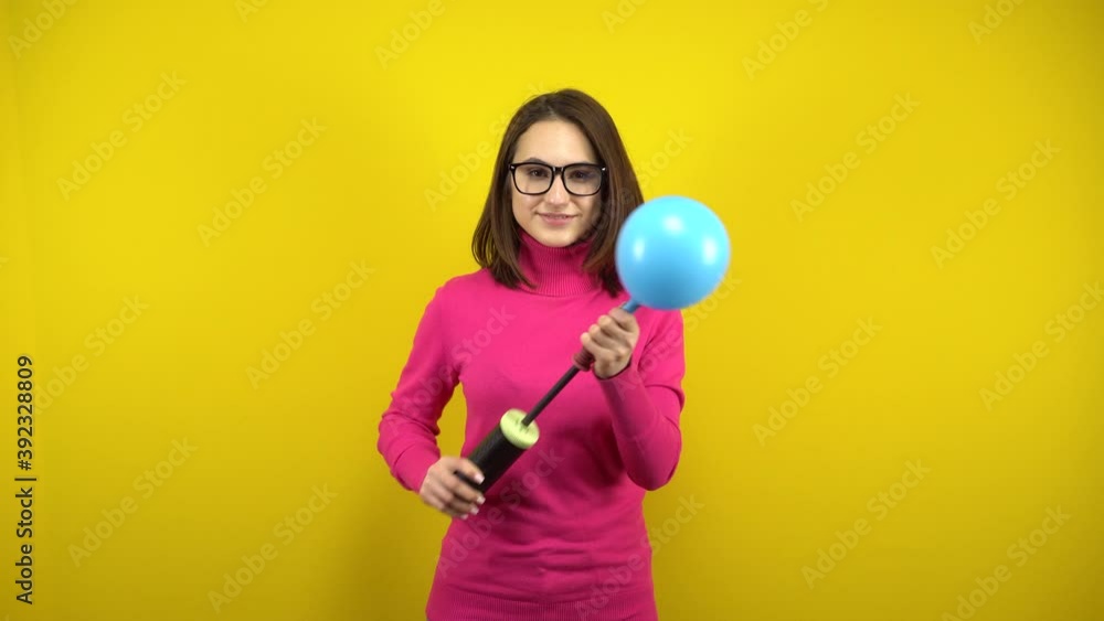 A young woman inflates a blue balloon with a pump on a yellow background. Girl in a pink turtleneck and glasses. A young woman inflates a blue balloon with a pump on a yellow background. Girl in a pink turtleneck and glasses.