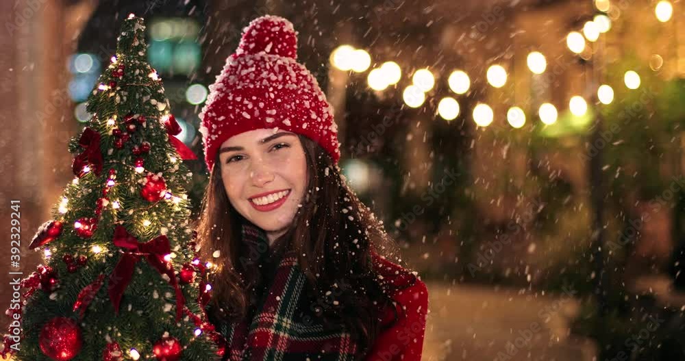 Joyful Caucasian young woman in good mood standing on street with small new years tree, looking at camera and smiling Happy female in christmassy town at night Close up portrait concept Joyful Caucasian young woman in good mood standing on street with small new years tree, looking at camera and smiling Happy female in christmassy town at night Close up portrait concept
