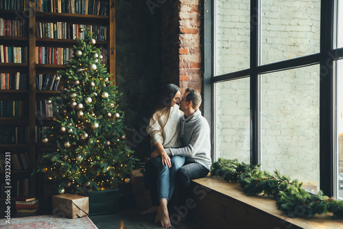 Couple in love near festive Christmas tree. A beautiful young wife and her husband sit on the floor in tenderly embrace. New Year eve together