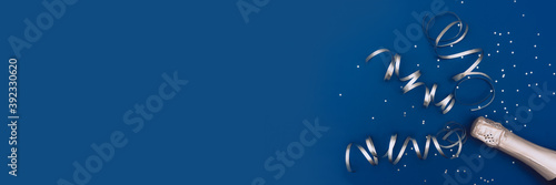 Photo banner of champagne bottle with confetti stars and party streamers on trendy classic blue background. Christmas, birthday or wedding concept. Flat lay, top view, copy space, mock-up.