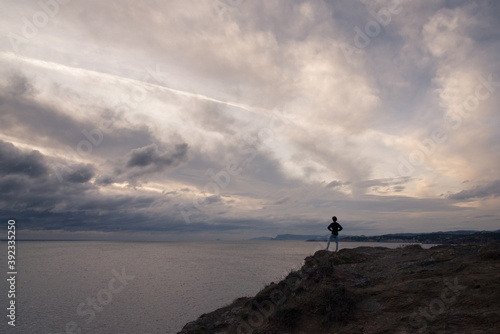 une personne de dos regarde la mer. Un ciel orageux sur la mer. Une personne sur un   peron rocheux regarde la mer.