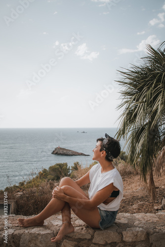 Beautiful woman with short hair looking at the sea smiling while sitting over a cliff in Ibiza wearing denim shorts