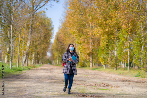 new normal Autumn walk outdoors - young happy and pretty Asian Japanese woman in face mask walking cheerful at beautiful city park in vibrant yellow and orange tree leaves © TheVisualsYouNeed
