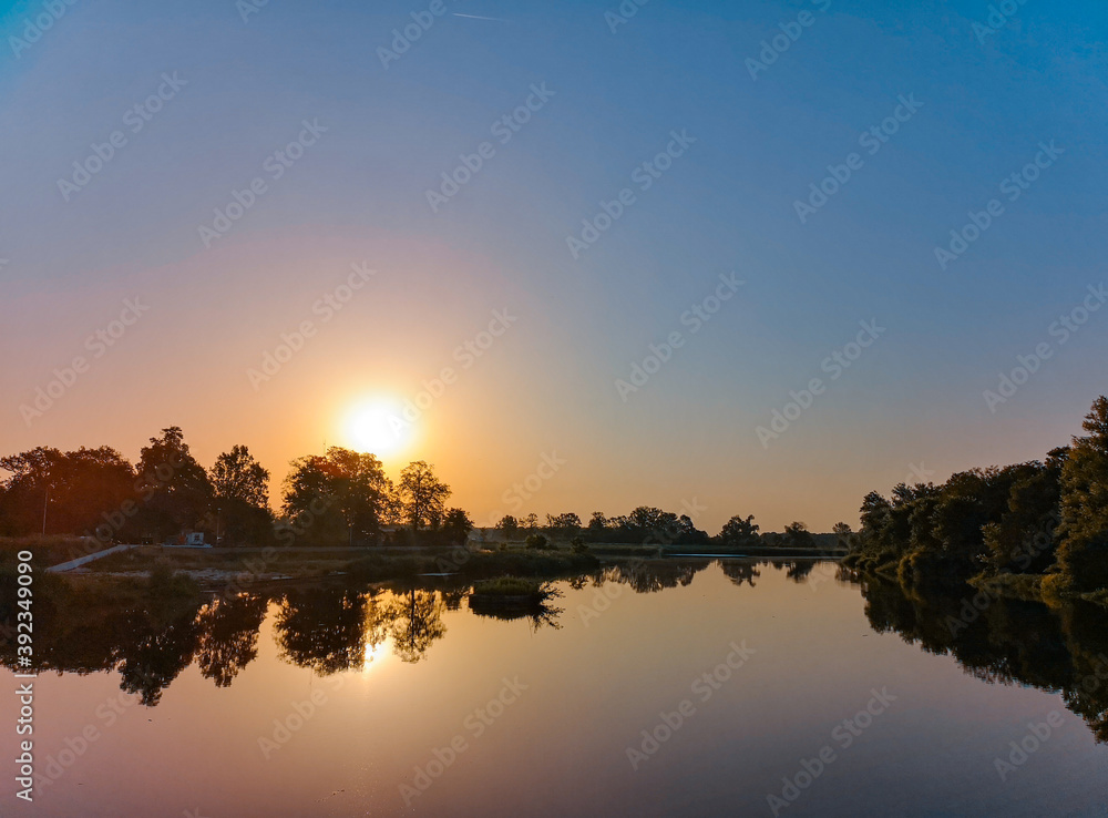 Fototapeta premium Rising sun over long river with waves between trees at sunny morning