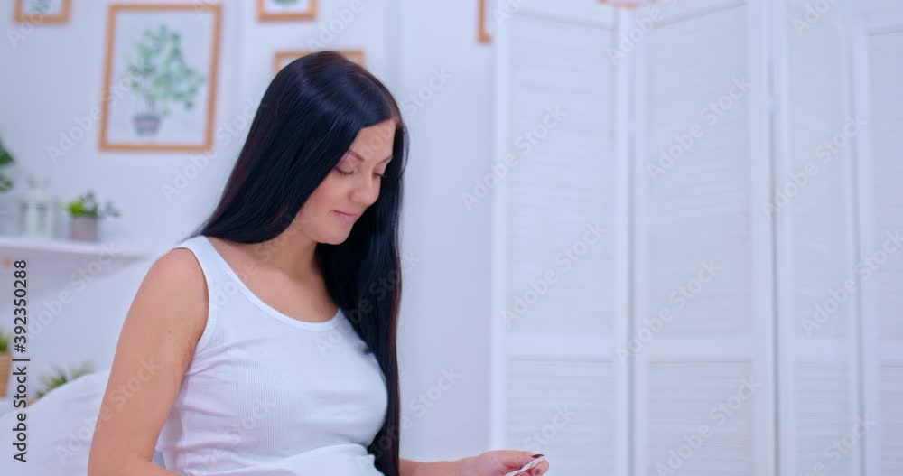 Portrait view, a beautiful young pregnant woman looks at a picture of a baby and strokes a tummy. Pink booties are on the bed, family values. 4k, ProRes Portrait view, a beautiful young pregnant woman looks at a picture of a baby and strokes a tummy. Pink booties are on the bed, family values. 4k, ProRes