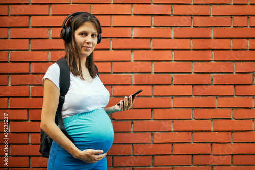 Pregnant sportswoman listening music and texting while standing outdoors. © chika_milan