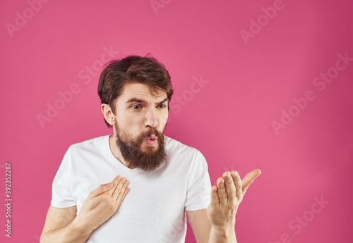 An indignant man gestures with his hands in a white T-shirt on a pink background