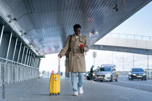 Portrait of smiling Afro-American millennial traveler man walking with yellow suitcase in airport terminal after arrival or to boarding gate, using mobile smart phone, chatting with friends. 