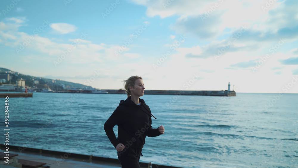 A young blonde girl in the early morning is engaged in jogging along the sea against the background of the lighthouse.