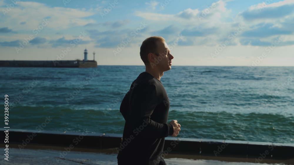 A young guy in the early morning is engaged in jogging along the sea against the background of the lighthouse.