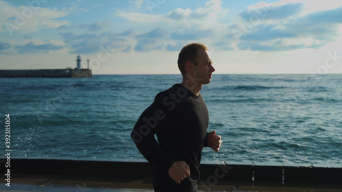 A young guy in the early morning is engaged in jogging along the sea against the background of the lighthouse.