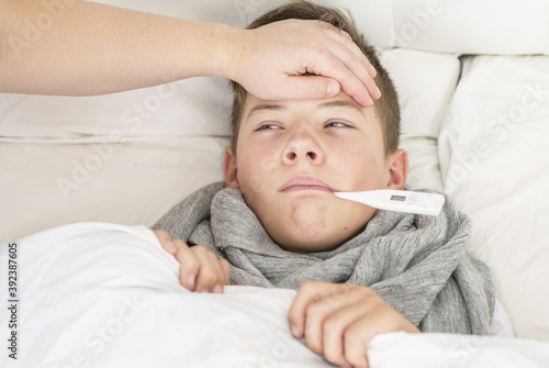 Fototapeta Sick  boy lies on bed during illness at home with a thermometer in his mouth