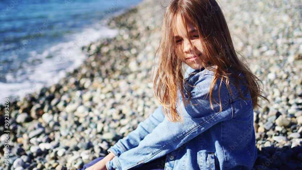 A beautiful teenage girl in a denim jacket sits on a pebble seashore A beautiful teenage girl in a denim jacket sits on a pebble seashore