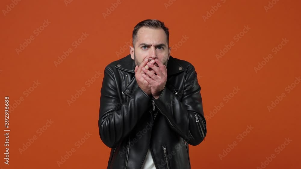 Shocked worried scared young bearded man 20s years old wearing black leather jacket isolated on orange color background in studio. People lifestyle concept. Looking camera covering mouth with hands