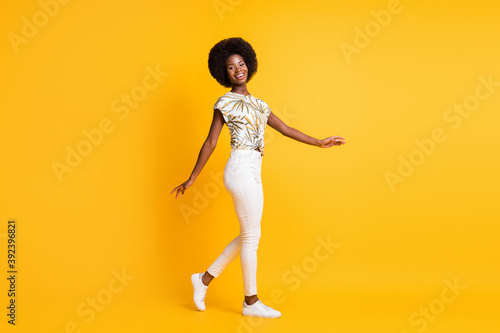 Full size profile photo of optimistic nice curly hair lady walking dress bright t-shirt sneakers trousers isolated on yellow background