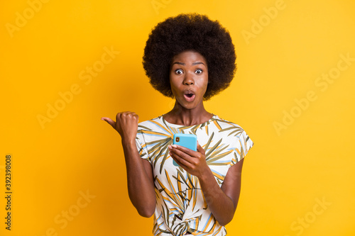 Photo of impressed pretty curly hair lady typing telephone point empty space wear print t-shirt isolated over yellow color background