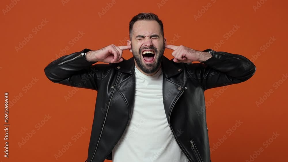 Crazy displeased young bearded man 20s in black leather jacket posing isolated on orange color background studio. People lifestyle concept. Covering ears with fingers screaming keeping eyes closed Crazy displeased young bearded man 20s in black leather jacket posing isolated on orange color background studio. People lifestyle concept. Covering ears with fingers screaming keeping eyes closed