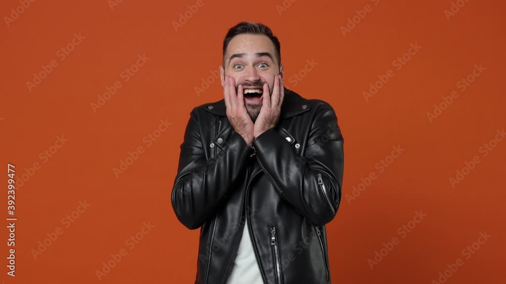 Side profile view surprised young man 20s in black leather jacket isolated on orange color background studio. People lifestyle concept. Turns around camera say wow put palms on cheeks spreading hands Side profile view surprised young man 20s in black leather jacket isolated on orange color background studio. People lifestyle concept. Turns around camera say wow put palms on cheeks spreading hands