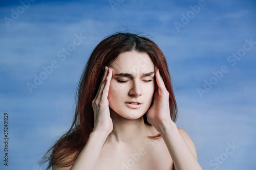 A woman with red hair twisted her face in pain and raises her hands to her face on blue background