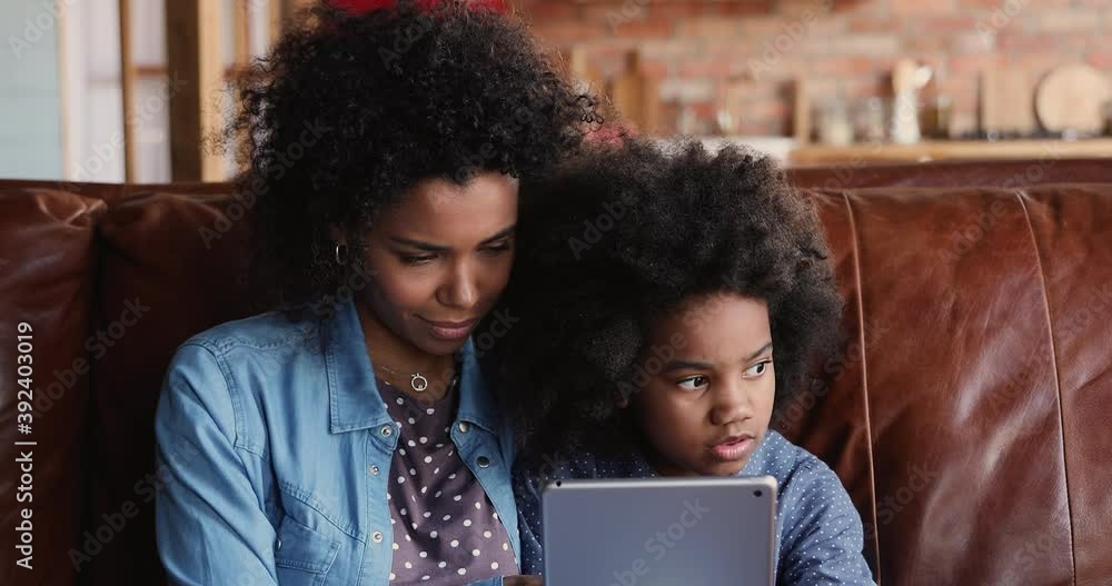 Close up view attractive African mother sit on couch with preschool kid girl, educate little daughter hold wireless modern tablet device using internet website online app for child development concept Close up view attractive African mother sit on couch with preschool kid girl, educate little daughter hold wireless modern tablet device using internet website online app for child development concept