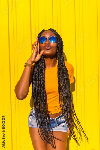 Lifestyle, pretty black girl with long braids, yellow t-shirts and short jeans on a yellow background. Smiling in sunglasses and having fun at photoshoot