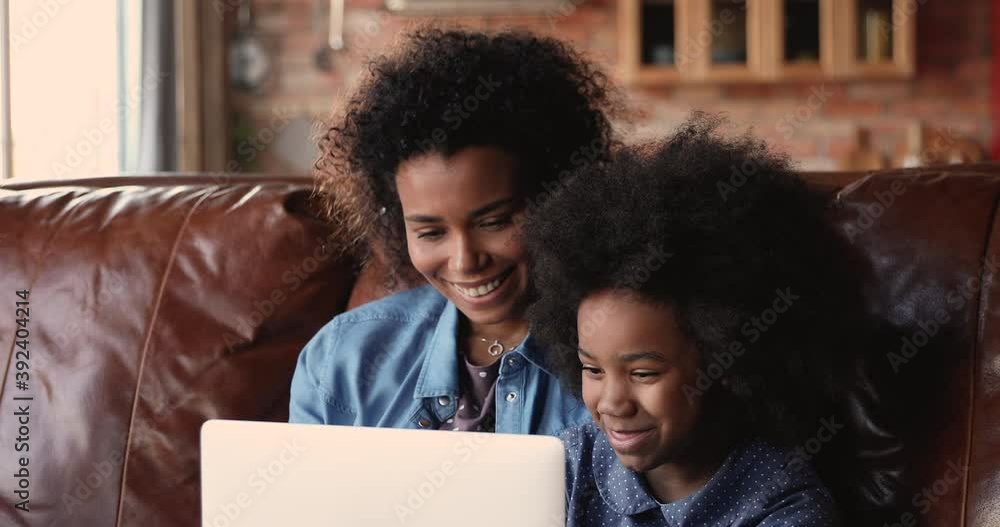 Happy African mom and little preschool daughter sitting on comfortable leather sofa in living room using laptop, having fun on internet, choose toys, buying on-line, play games, watch cartoons concept Happy African mom and little preschool daughter sitting on comfortable leather sofa in living room using laptop, having fun on internet, choose toys, buying on-line, play games, watch cartoons concept