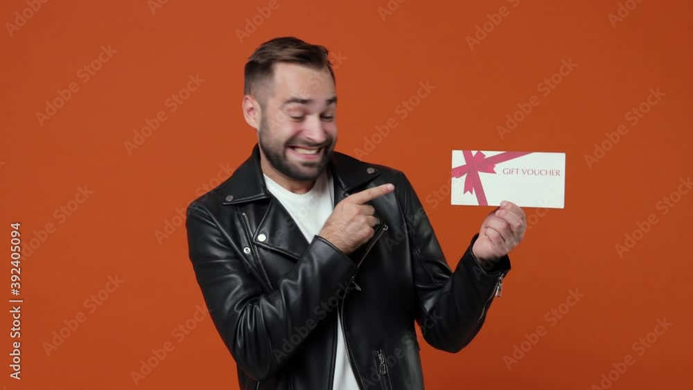 Excited young bearded man 20s in casual black leather jacket isolated on orange background in studio. People lifestyle concept. Pointing index finger on gift certificate camera doing winner gesture Excited young bearded man 20s in casual black leather jacket isolated on orange background in studio. People lifestyle concept. Pointing index finger on gift certificate camera doing winner gesture