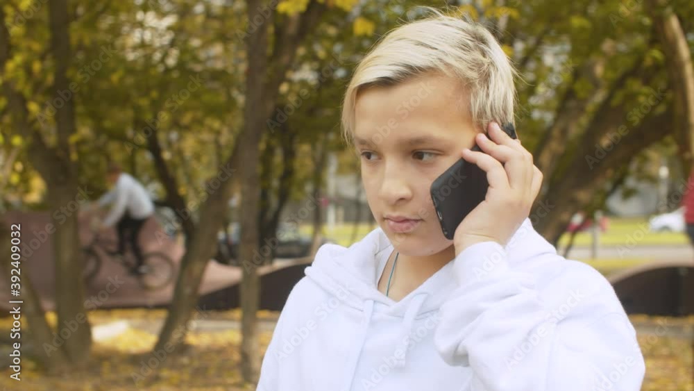 Portrait of blond teenager boy talking by smart phone in the park. Blurry unrecognizable children riding scooters on the pump track, summer day Portrait of blond teenager boy talking by smart phone in the park. Blurry unrecognizable children riding scooters on the pump track, summer day