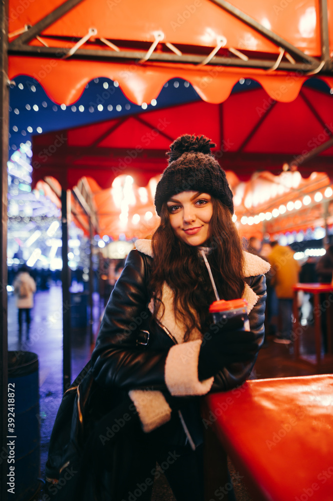 A positive lady in a hat stands at an evening fair with a cup of hot drink in her hand, looks into the camera and smiles, wears winter clothes. Walk on the Christmas market. A positive lady in a hat stands at an evening fair with a cup of hot drink in her hand, looks into the camera and smiles, wears winter clothes. Walk on the Christmas market.