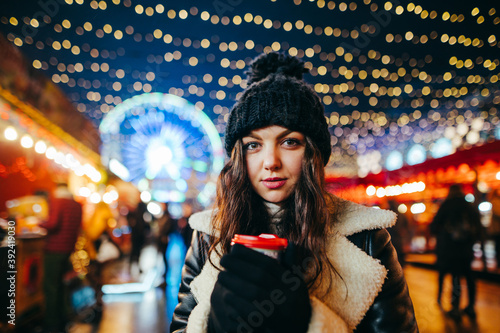 Portrait of a pretty girl in warm clothes standing at a Christmas party with a cup of hot drink in her hands, looking at the camera, close photo. Christmas portrait of a cute lady at the fair.