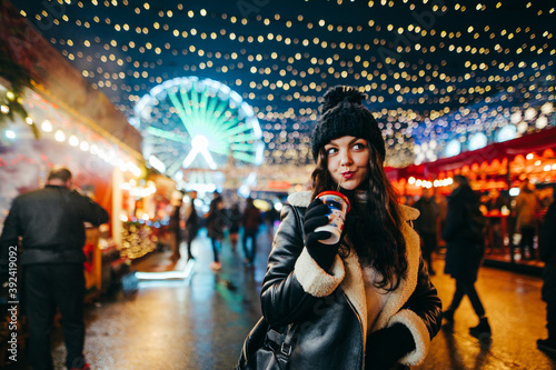 Evening portrait of a cute girl in warm clothes stands at a decorated Christmas market and drinks warming drinks.Lady walks down the street at Christmas and drinks a hot drink on a bright background.