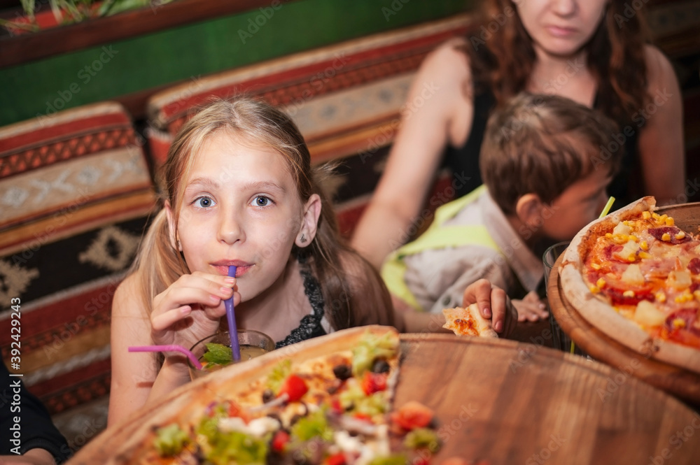 A young girl with her family is resting in a cafe, they are eating pizza and talking, the girl is drinking a soft drink with lemon, lime and mint through a straw