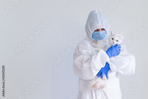 A woman in a protective suit with gloves and a mask is holding a white cat. Doctor tests pets for coronavirus