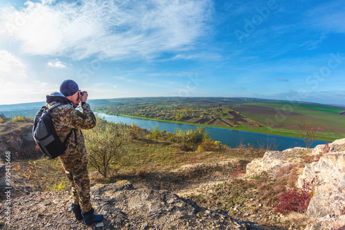 a man photographs the landscape near the river in camouflage clothes with a backpack on his back