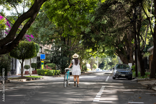 Mujer con vestido y sombrero caminando por la calle llevando una bicicleta, con arboles a los lados en un dia de invierno.